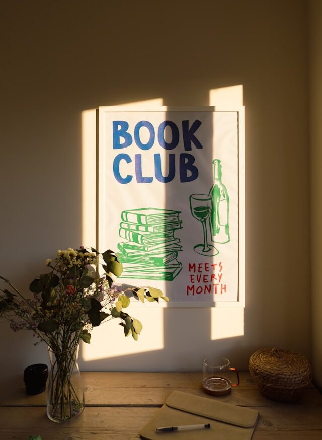 Foto de stock en donde se observa una mesa con flores y una copa de licor, y, al fondo, un cartel de un Club de Lectura.