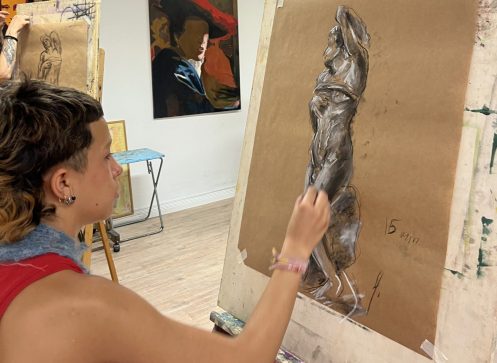 Joven de lado haciendo un cuadro con carboncillo encima de un caballete dentro de una escuela de arte.